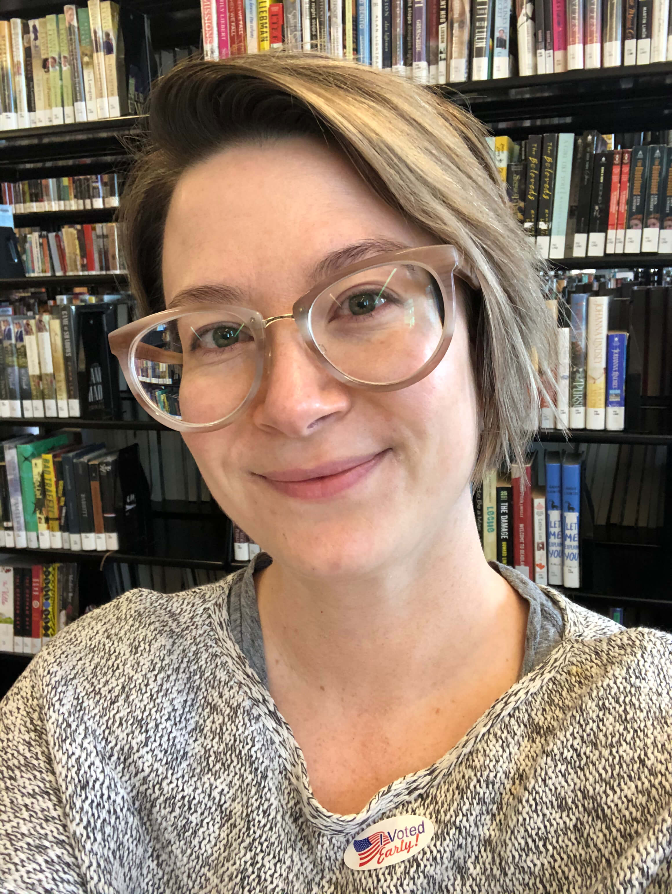 A photo of Sarah Rigdon, with library books behind her. She's wearing pink-rimmed glasses and a grey shirt with an "I Voted Early!" sticker on it.